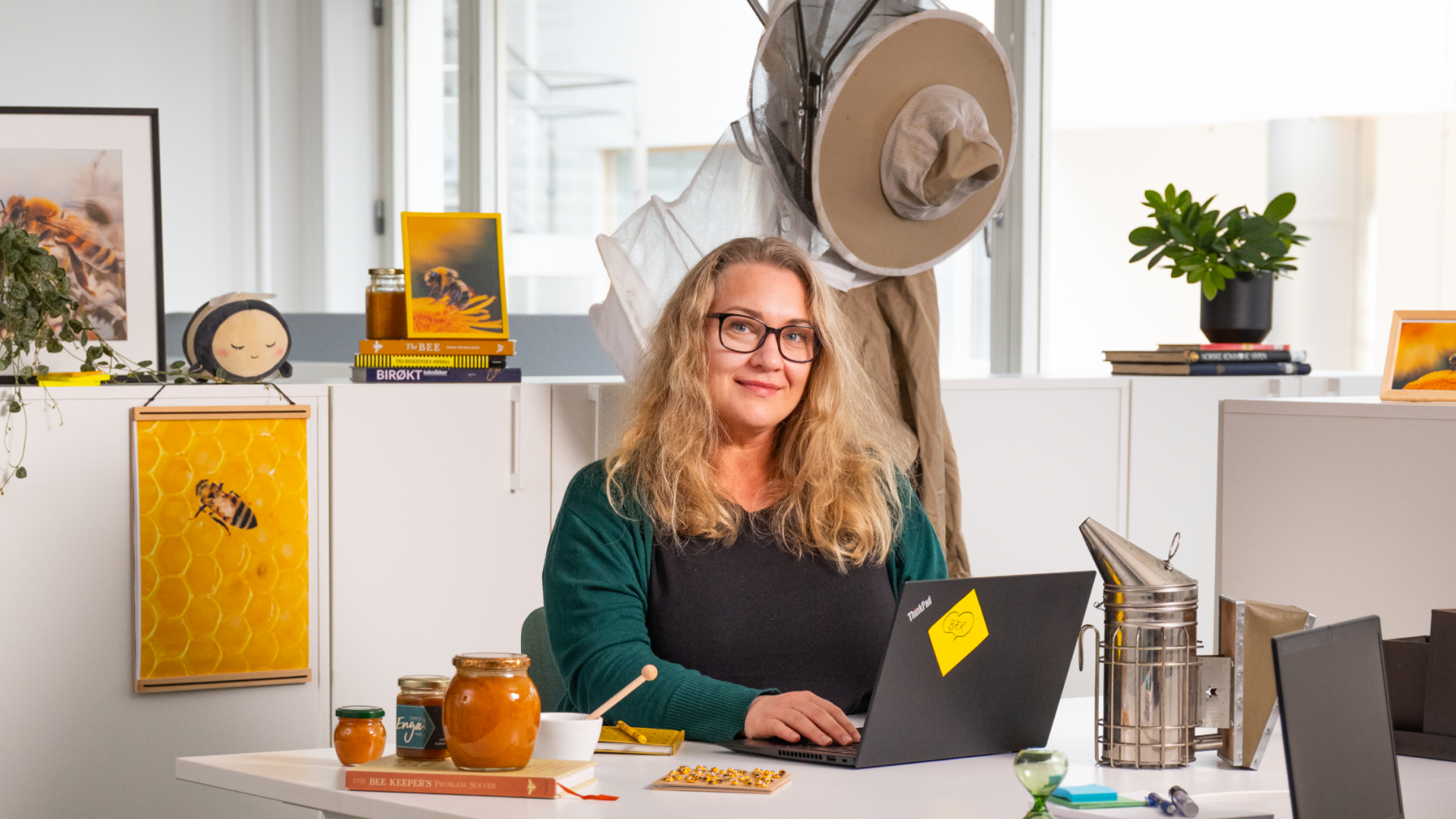 En dame sitter i et kontorlandskap foran en PC og smiler mot kamera